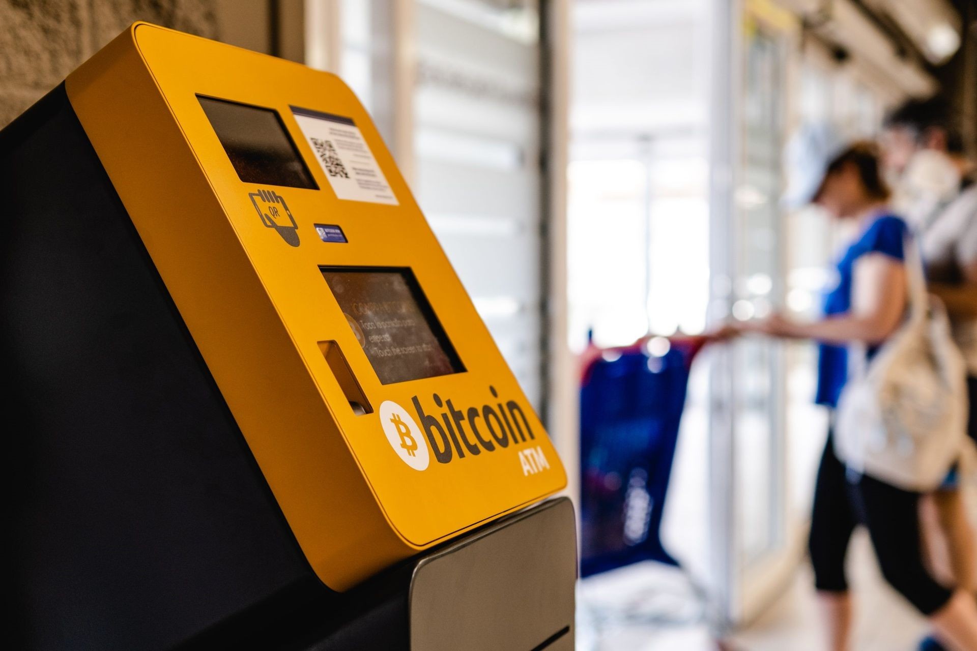 Close-up of a yellow Bitcoin ATM in a public area with blurred people in the background.